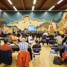 Room full of people sitting at tables for a luncheon and listening to a speaker
